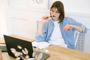 A young woman sits at a table looking at an open laptop and clicking a pen against her lip.